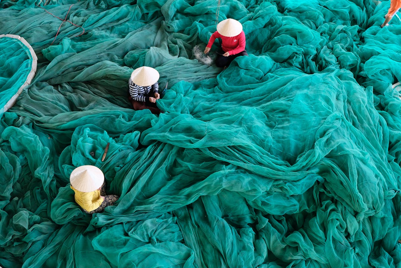 Aerial view of three people weaving turquoise fishing nets, showcasing traditional craftsmanship and textiles.