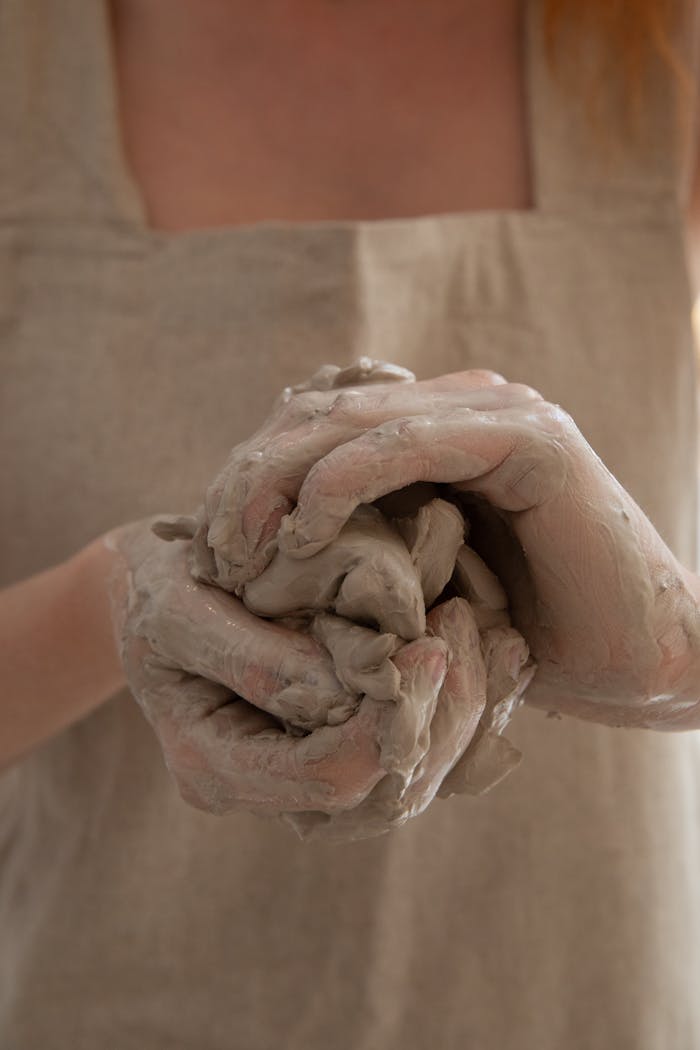 Unrecognizable person wearing gray apron shaping and molding clay in dirty hands while making handicraft crockery in workshop during work